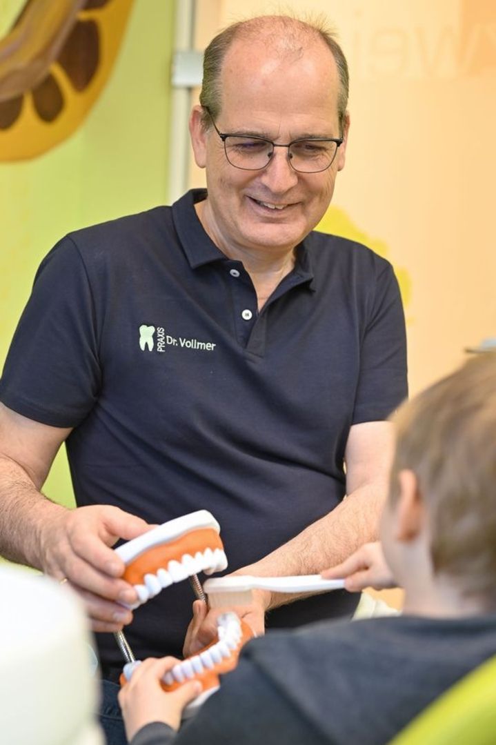 Ein Mann in einer Zahnarztpraxis zeigt einem Kind eine Prothese, während das Kind eine Zahnbürste hält.
